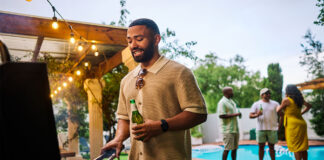 Man grilling food while holding a beer at an outdoor barbecue party by the pool. Group of friends socializing in the background. Summer celebration concept. Medium shot