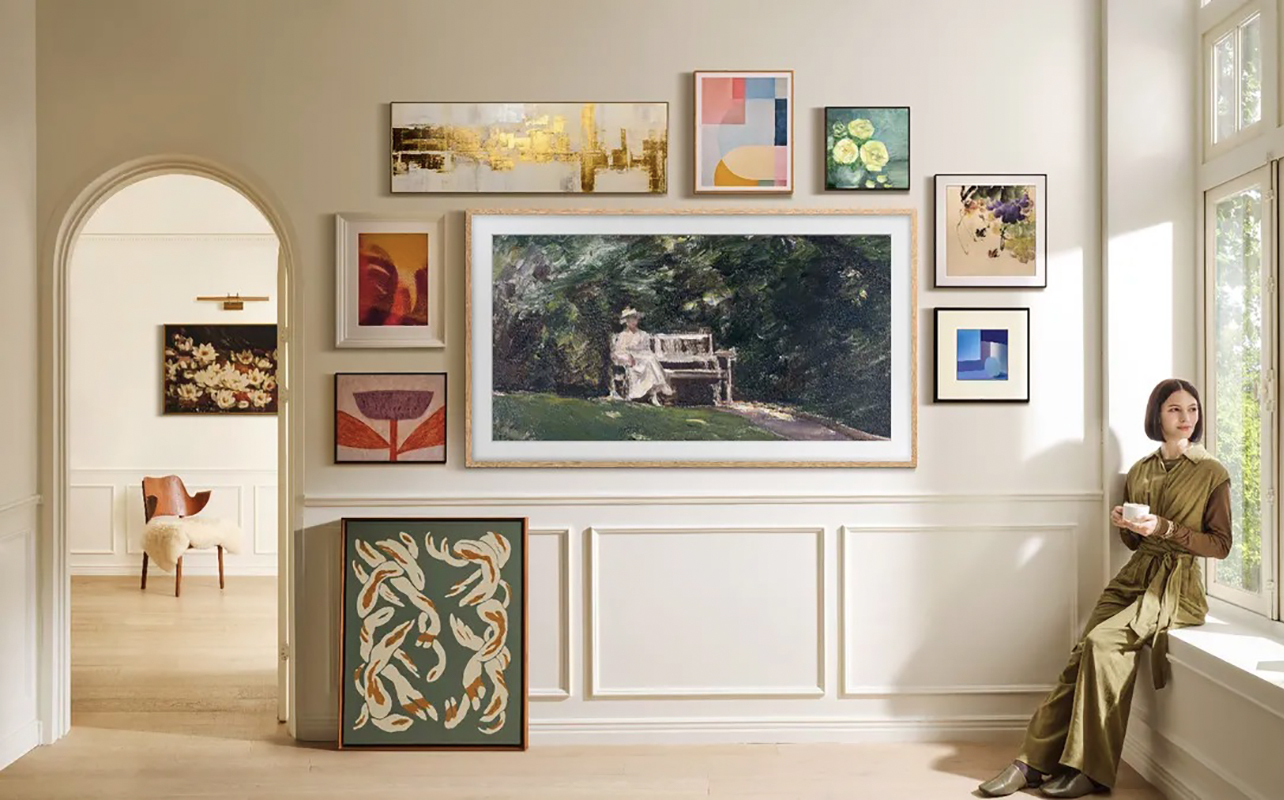 Un téléviseur The Frame de Samsung monté au mur avec des cadres photos plus petits autour, une femme assise sur le rebord de la fenêtre à droite.